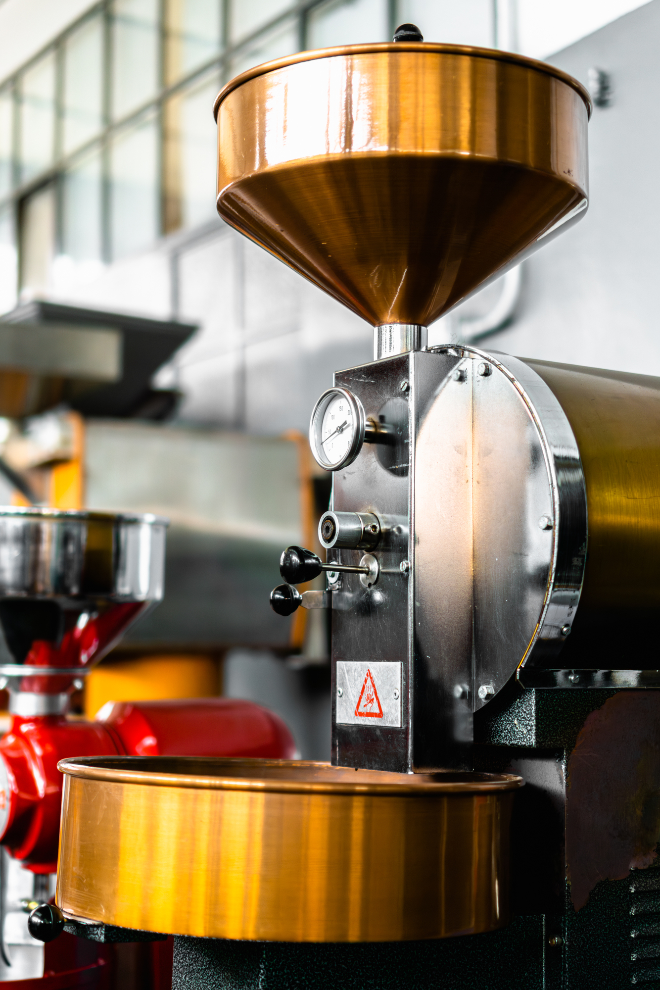 "Macchina per la torrefazione del caffè con tramogge in rame lucido e struttura in acciaio inox. La macchina è dotata di indicatori di temperatura, leve di regolazione e componenti meccanici di precisione. Sullo sfondo, altre attrezzature per la lavorazione del caffè in un ambiente industriale